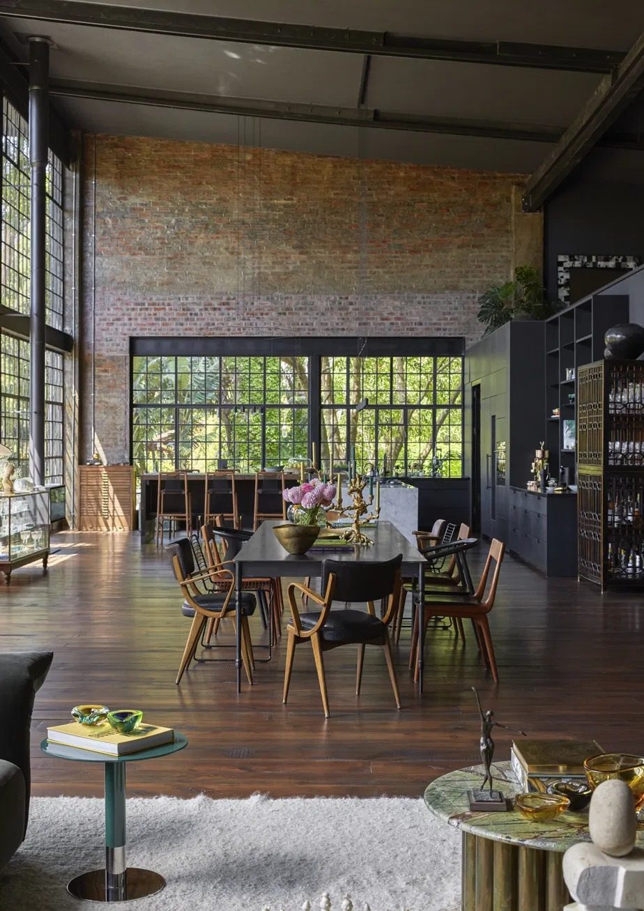 Long dining table with timber chairs in front of a brick wall and garden windows, with the kitchen and a bar cabinet to the right