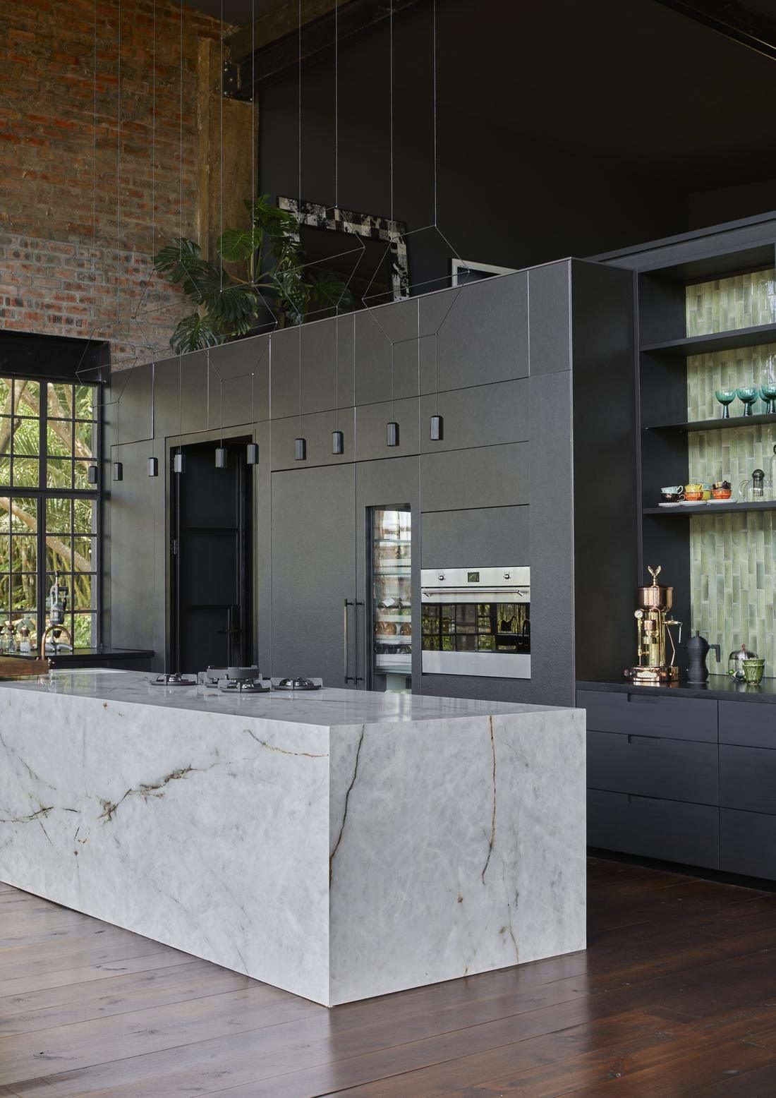 Marble kitchen island in front of a dark cabinet wall, with a copper espresso machine and green tiled shelves
