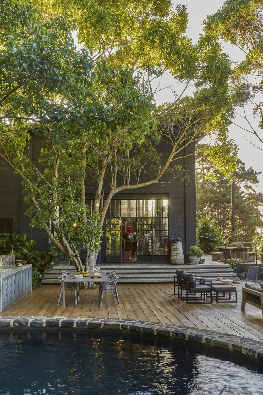 Timber deck with a dining table and lounge seating beside the black pool, the glass doors of the house glowing behind the trees