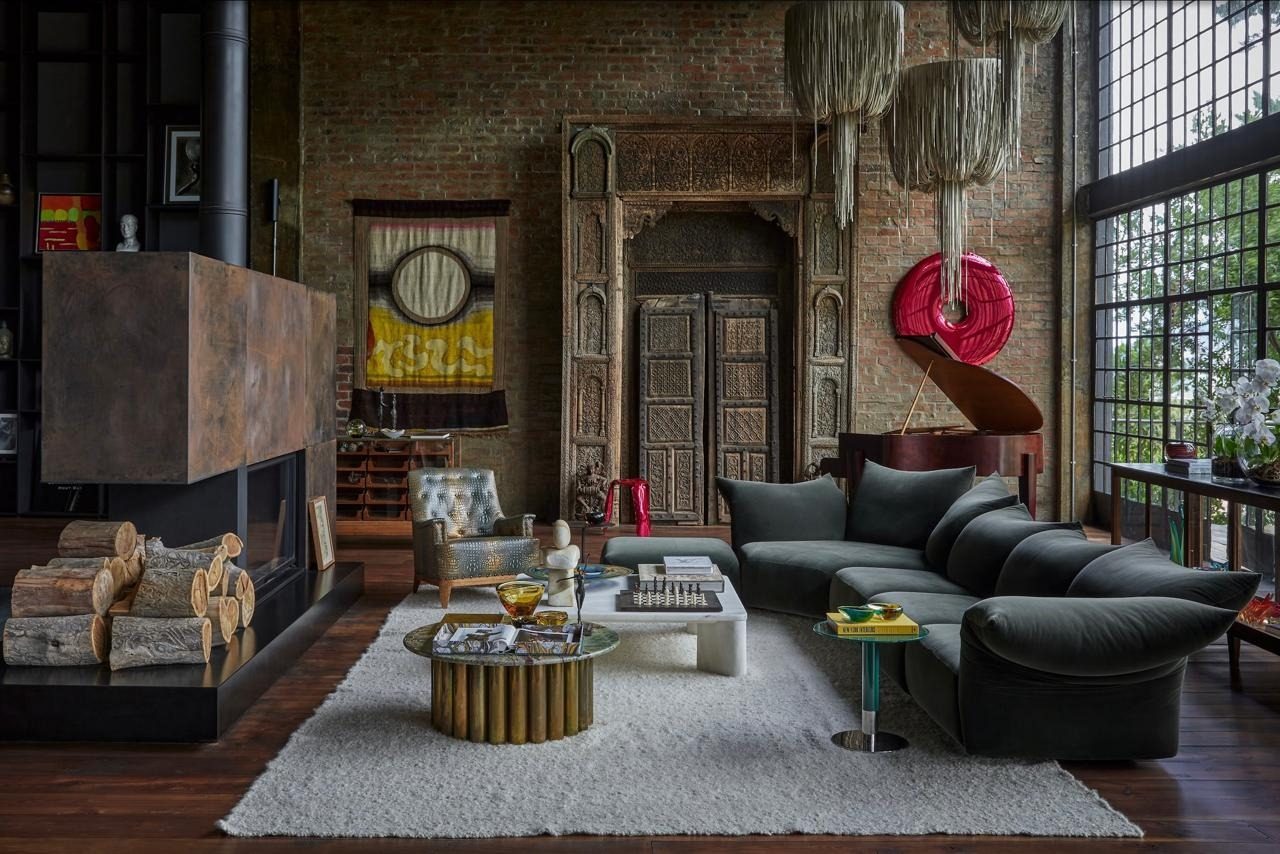 Double height living room with a brick wall, carved timber doorway, a fireplace with stacked logs, a green velvet sofa and a grand piano beneath a red sculpture